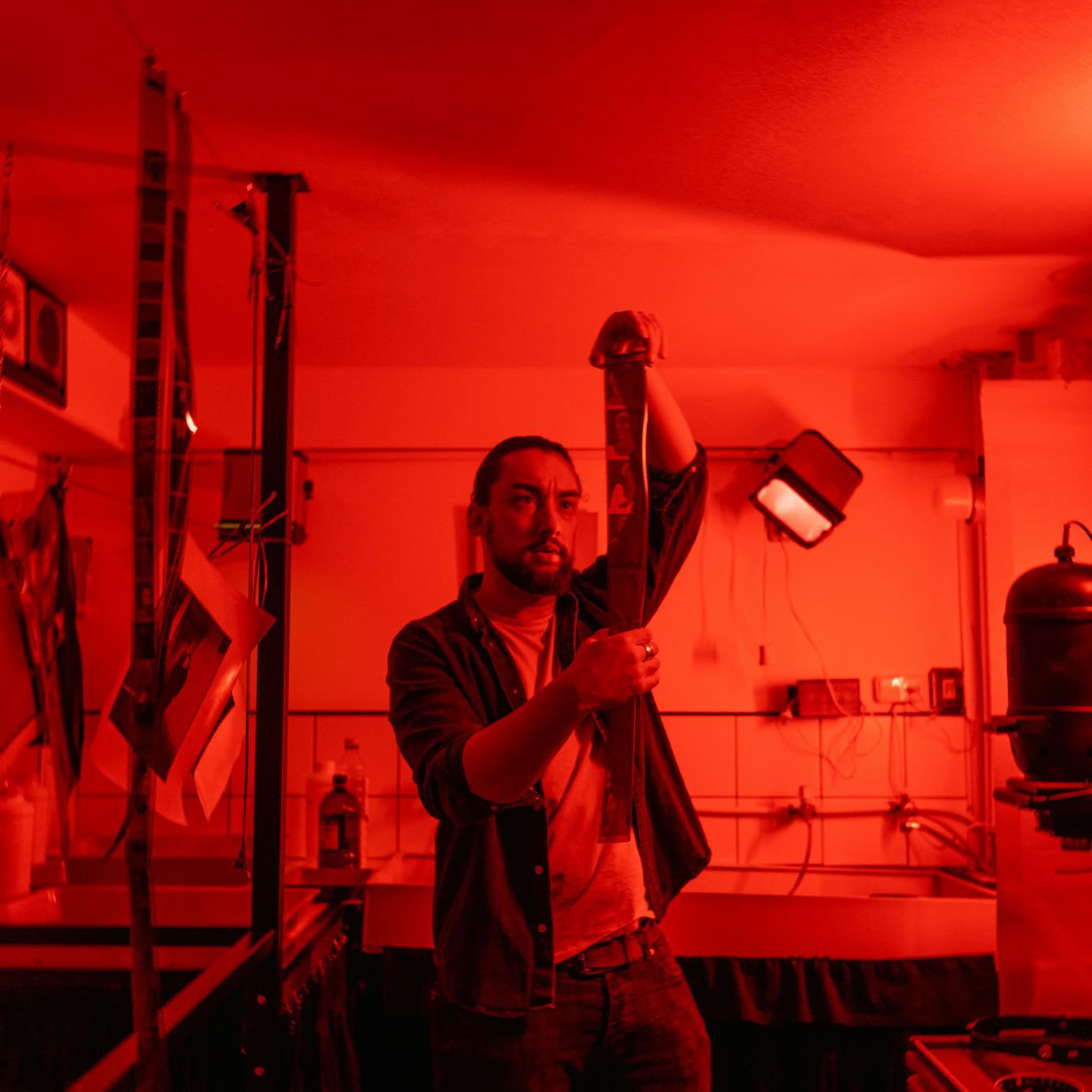 A photographer in a red-lit darkroom carefully examines a film strip during the developing process.